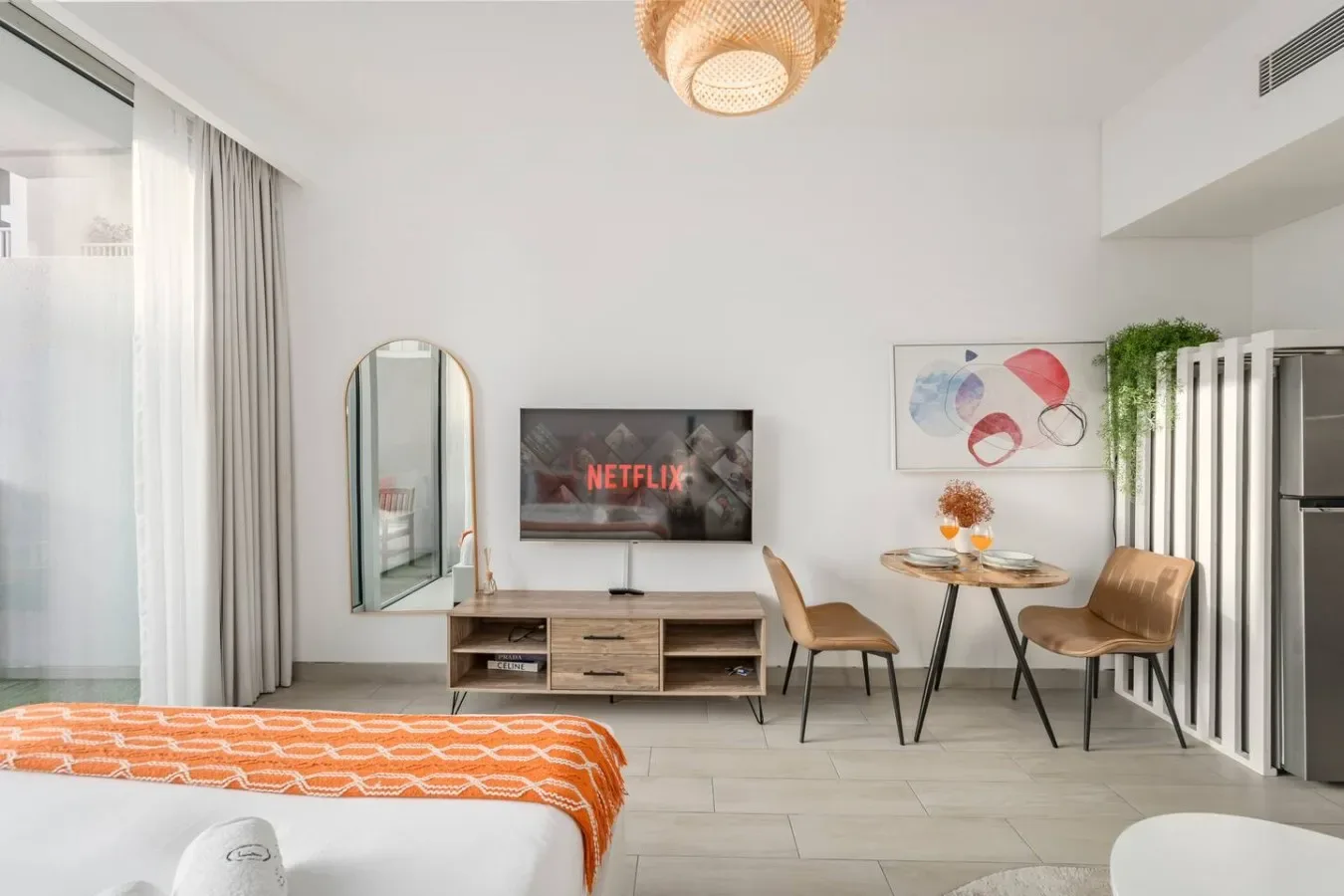Un dormitorio luminoso con televisor, una pequeña mesa de comedor y ropa de cama naranja estampada.