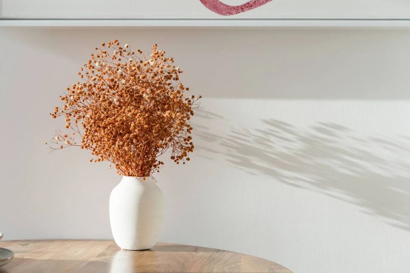 Un jarrón blanco con flores secas está sobre una mesa de madera.