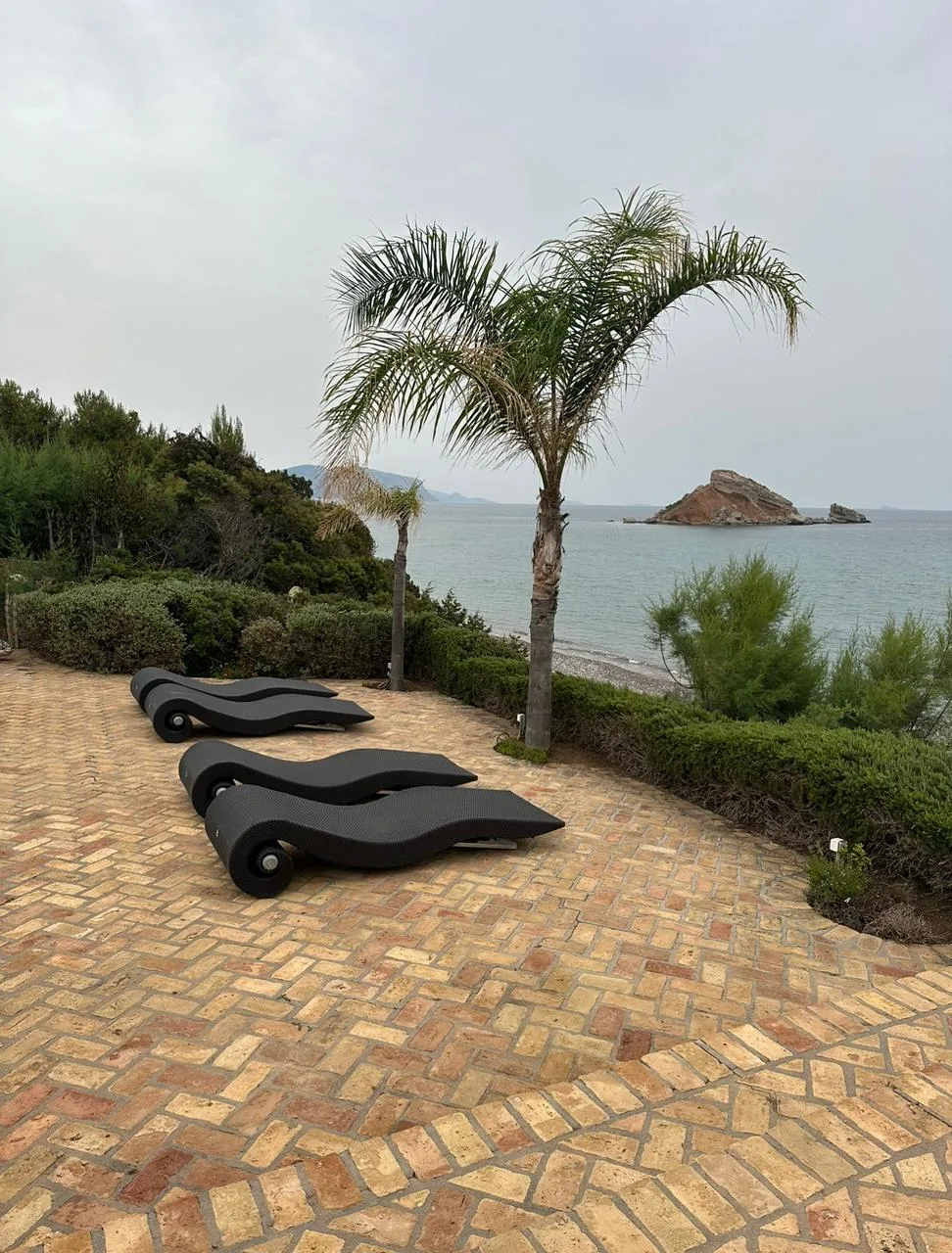 A brick-paved patio with lounge seating overlooks the sea and a rocky islet.
