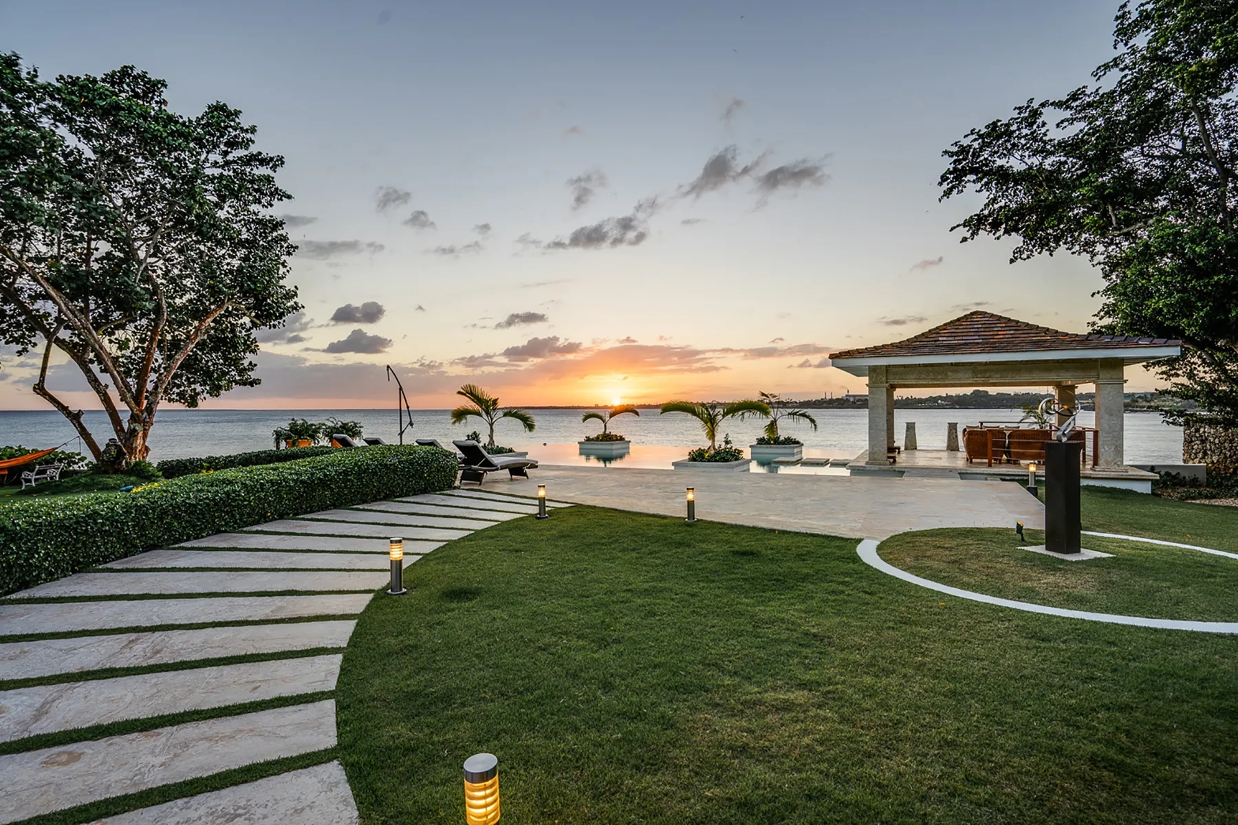 Un jardín junto al agua con un sendero pavimentado y el atardecer sobre el horizonte.