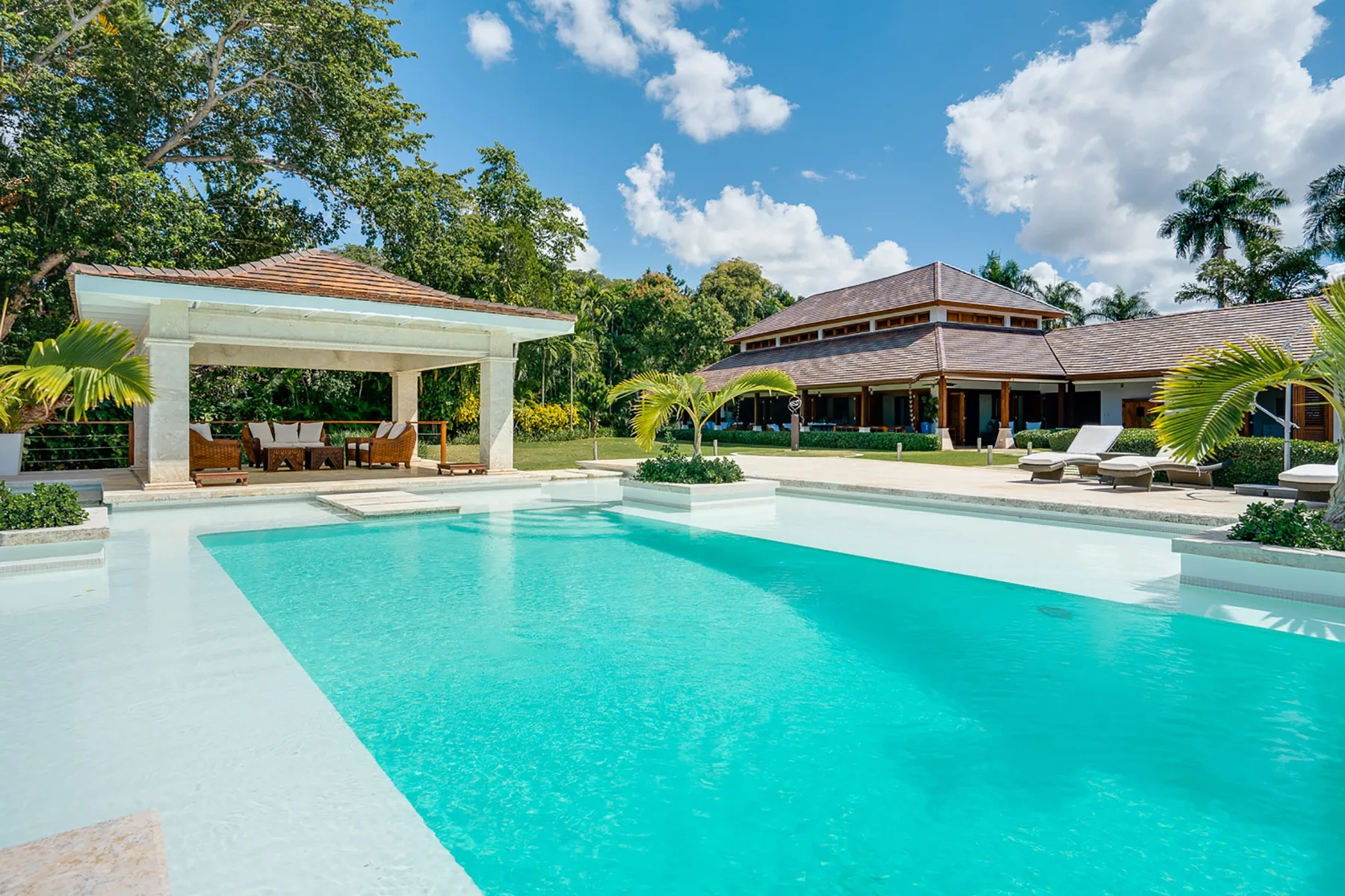 An outdoor swimming pool sits beside a covered seating area and low buildings.