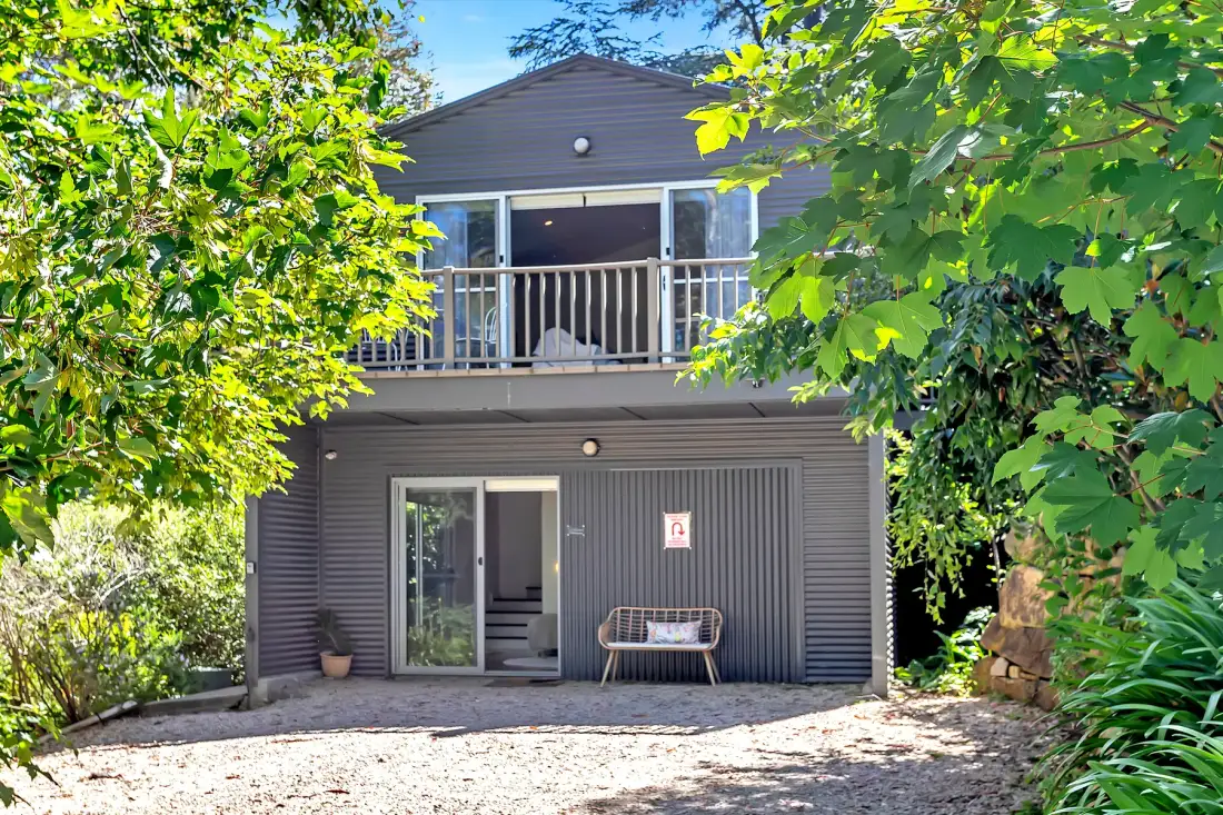 A two-story house stands among leafy trees, with a balcony above the ground-floor entrance.