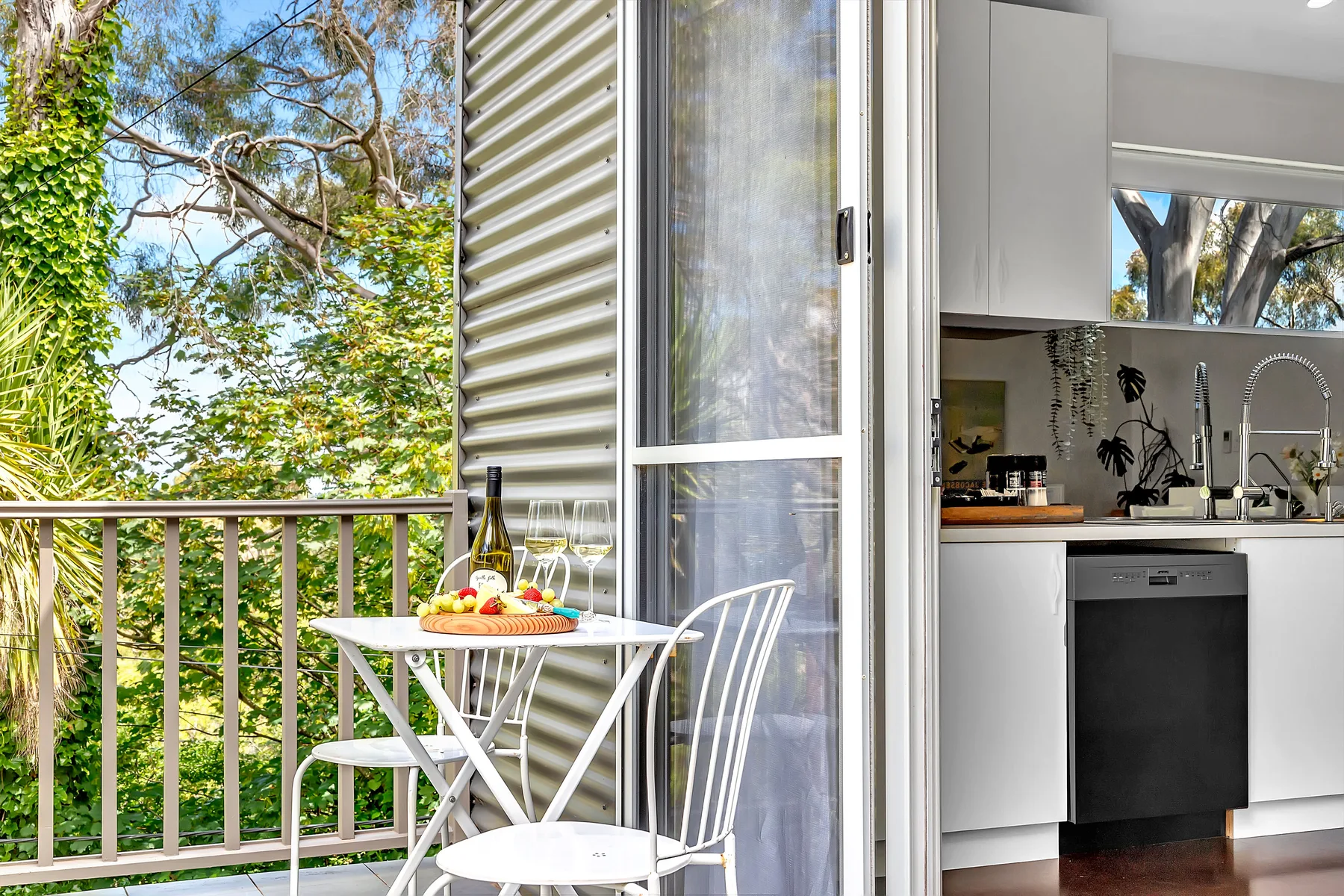 A small balcony with outdoor seating overlooks leafy trees beside an open kitchen.