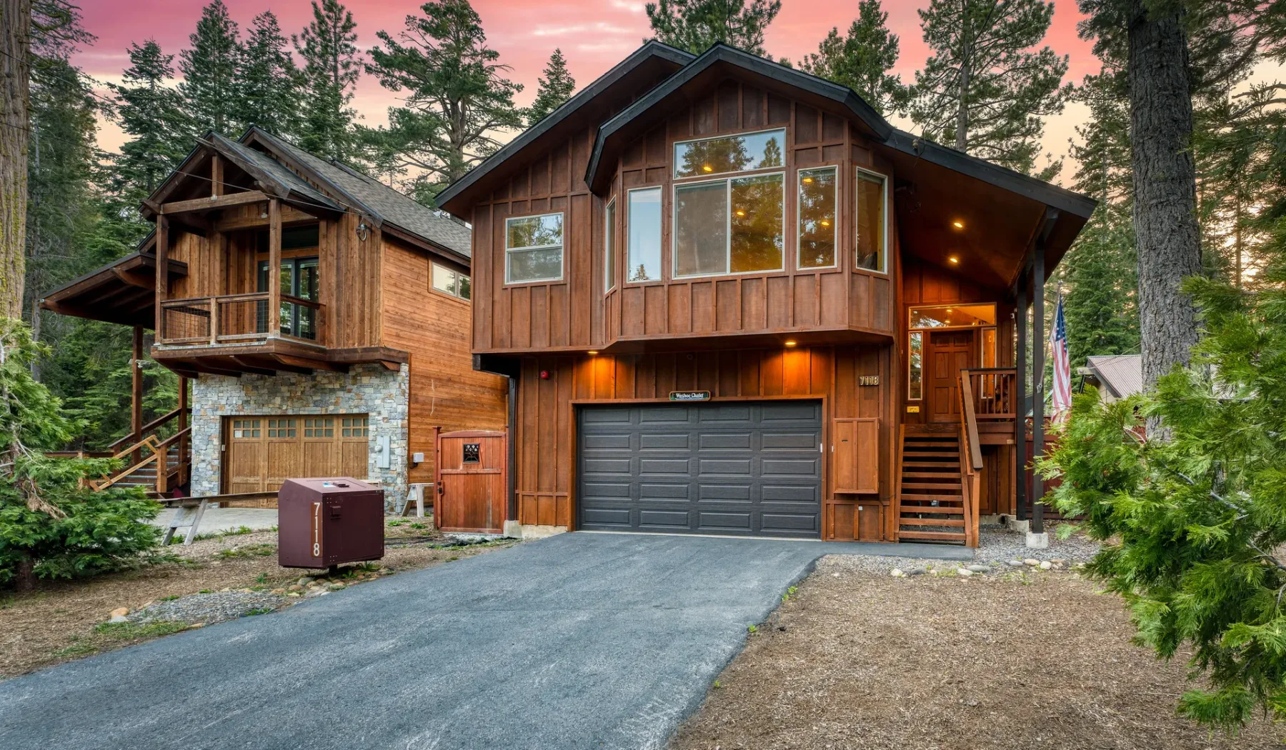 A large wooden house stands among tall pine trees beside a paved driveway.