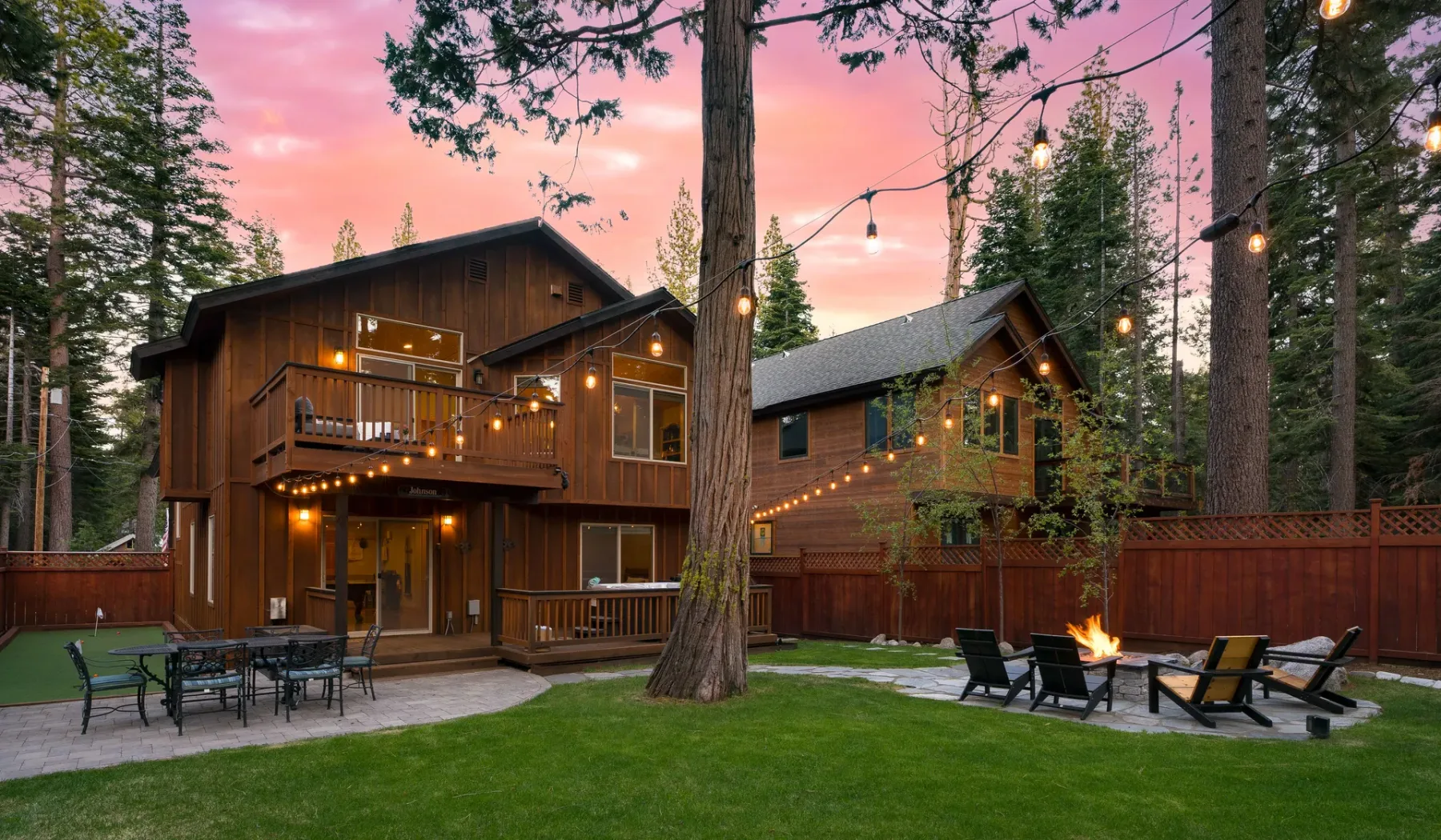 A wooded cabin courtyard with outdoor seating and a lit fire pit.