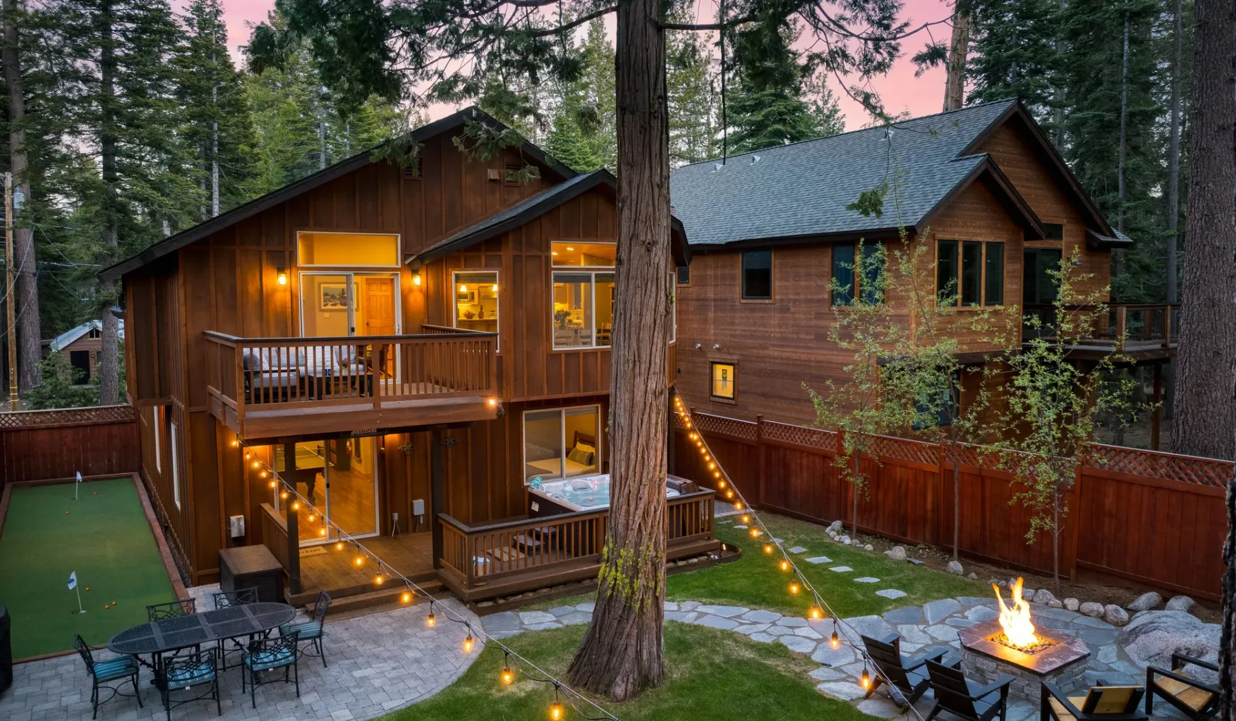 A wooden cabin with lit windows, outdoor seating, and a small fire pit among trees.