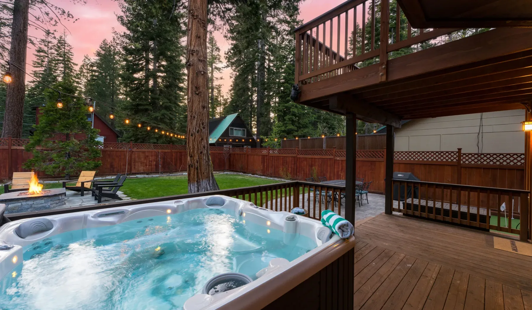 An outdoor deck features a bubbling hot tub beside a fenced yard and string lights.