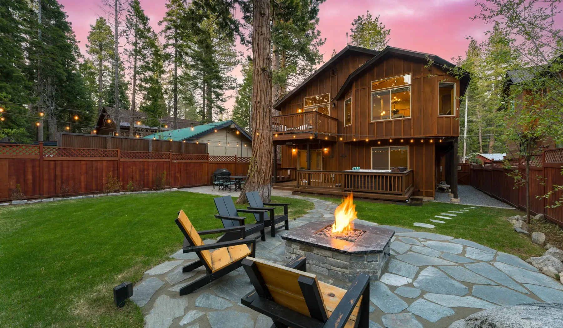 A wooden cabin with a deck overlooks a stone patio and lawn among trees.