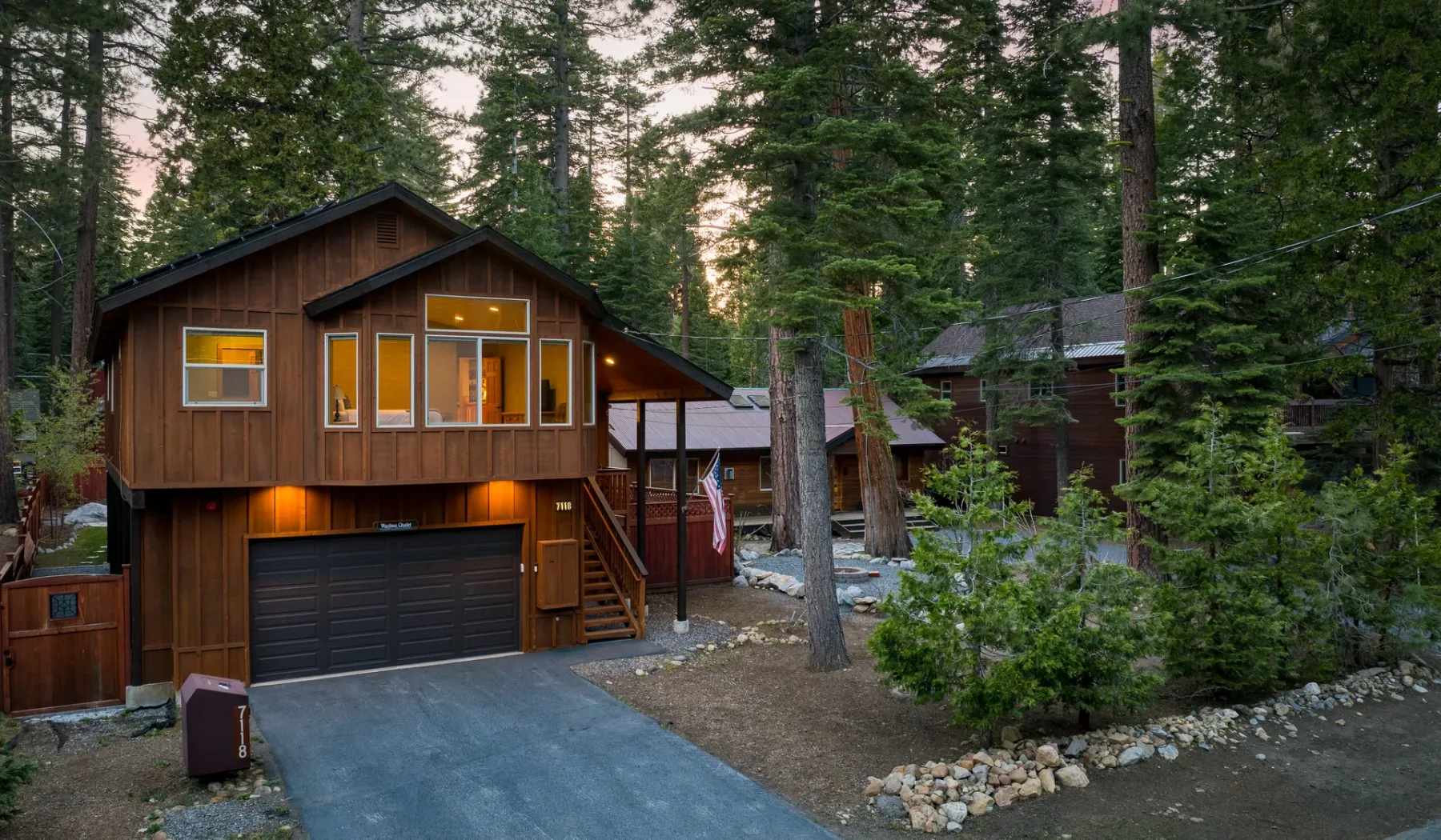 A wooden cabin sits among tall evergreen trees, with a driveway leading to its garage.