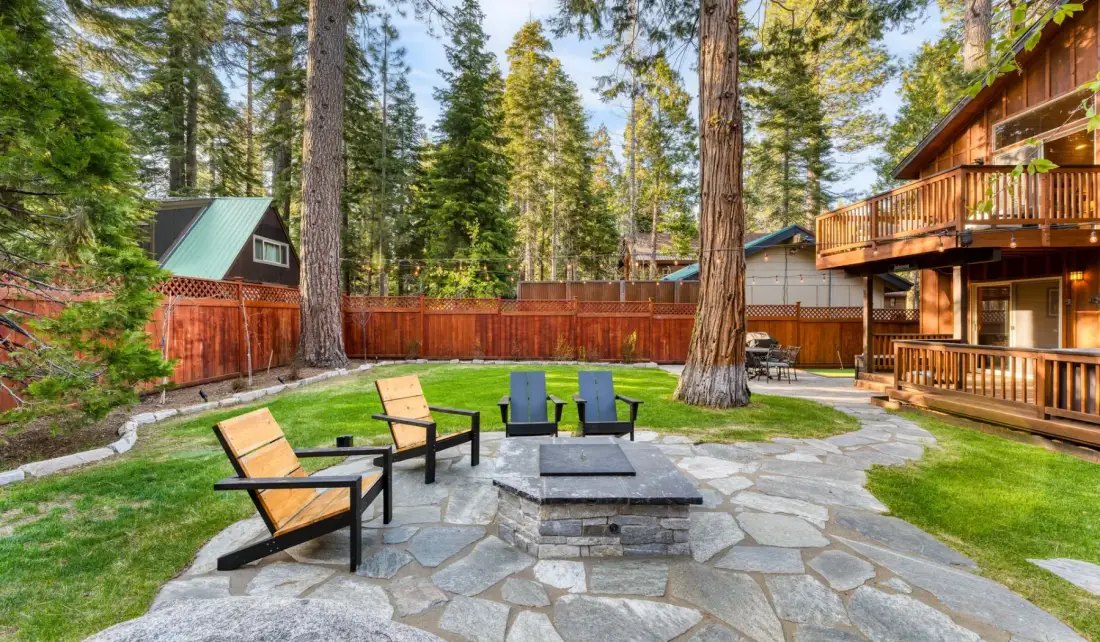 A stone patio with outdoor seating and a fire pit sits among tall trees beside a wooden cabin.