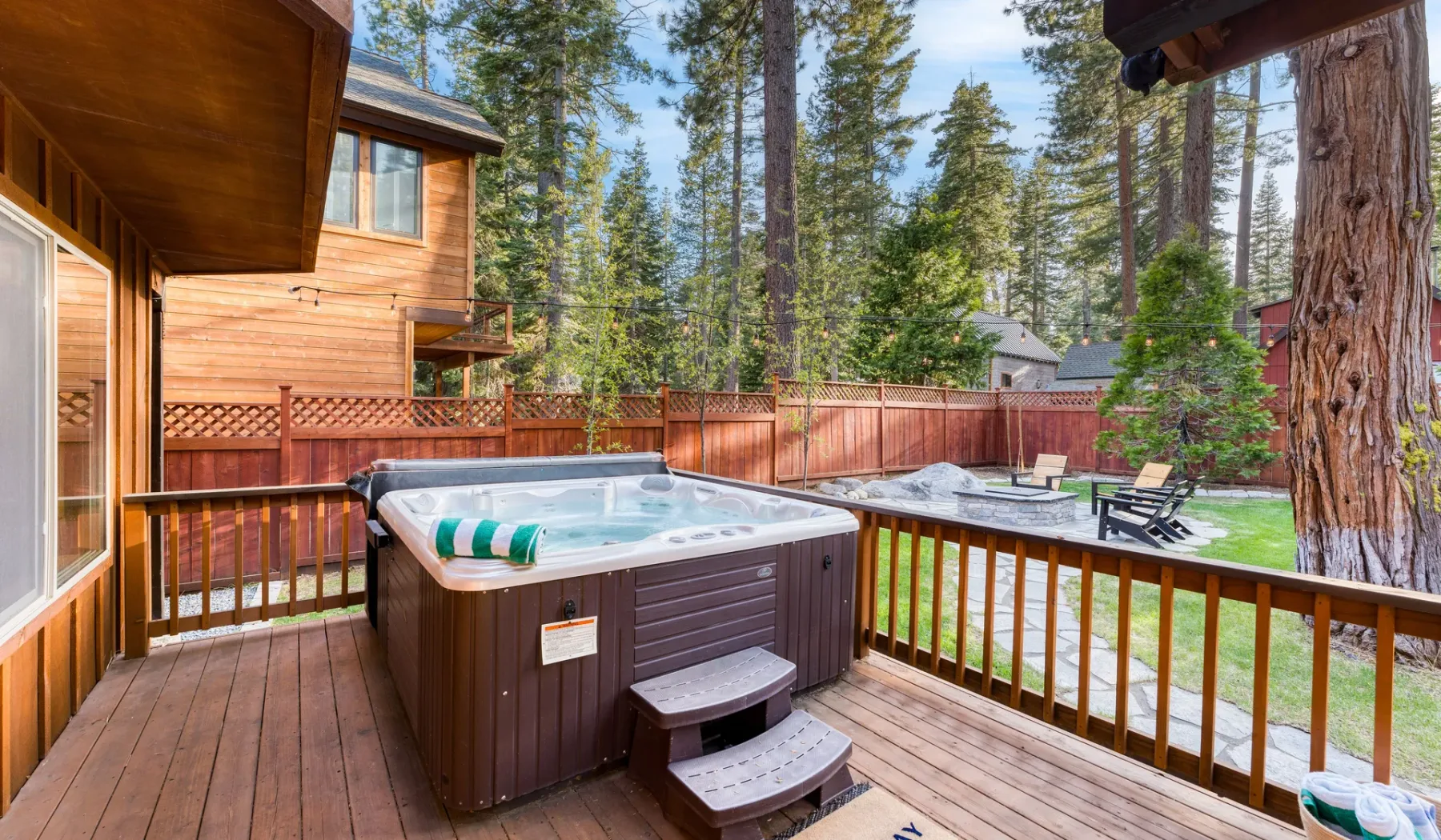 A wooden deck with a hot tub overlooks a fenced yard and surrounding trees.