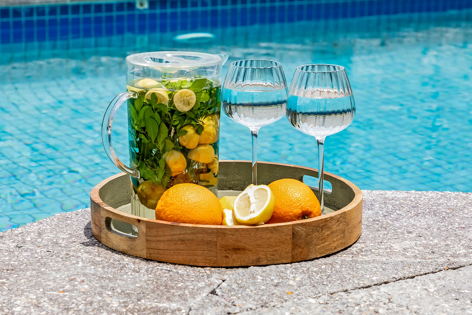 A wooden tray with citrus drinks and glasses sits beside a swimming pool.