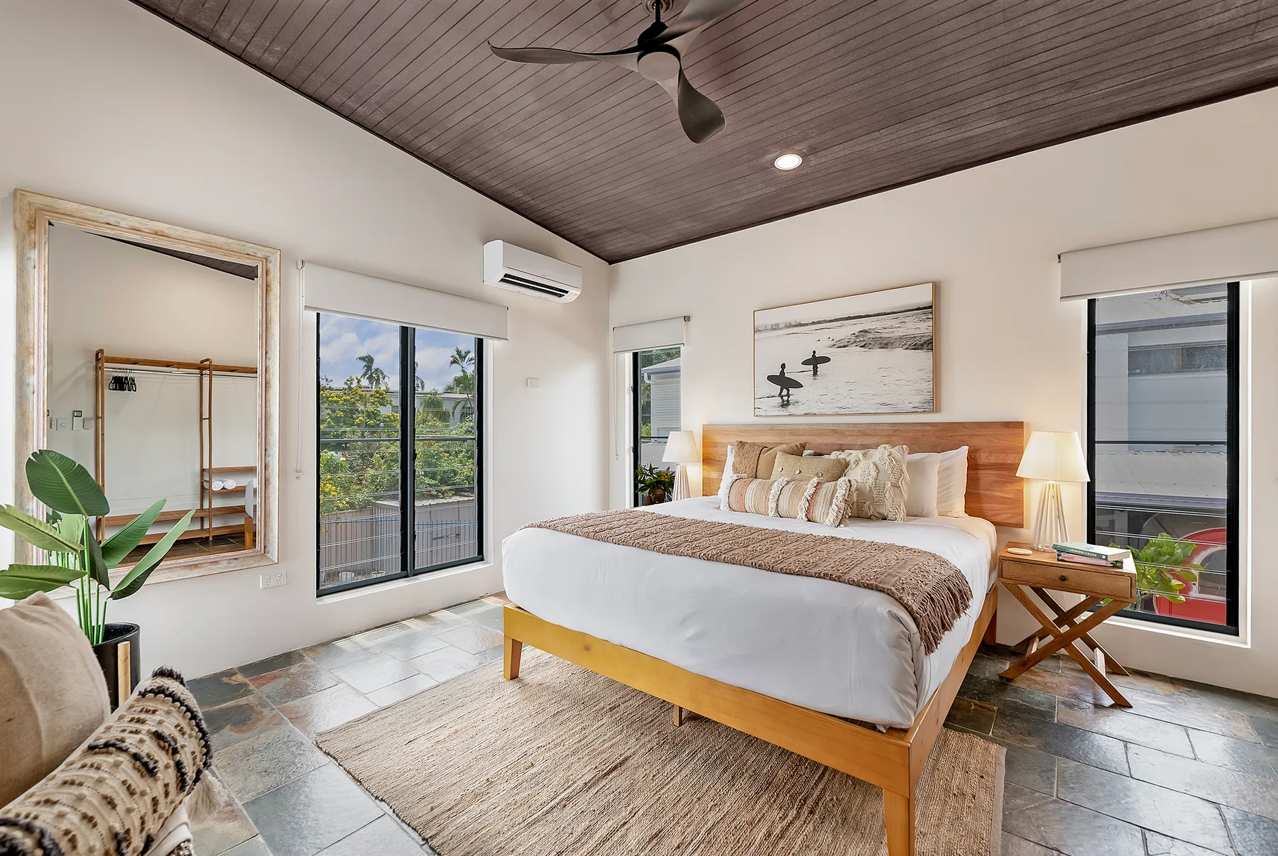 A bedroom with a large bed, ceiling fan, and windows overlooking greenery.