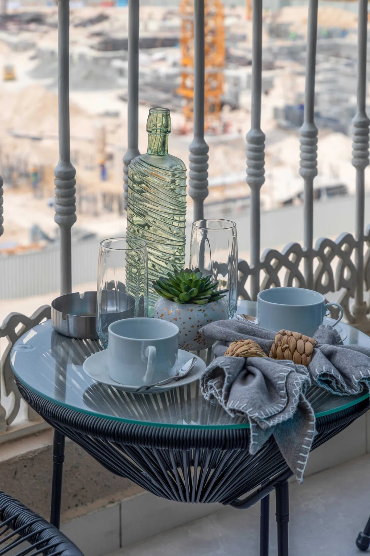 A small round table holds cups, glassware, a plant, and folded textiles by a window.