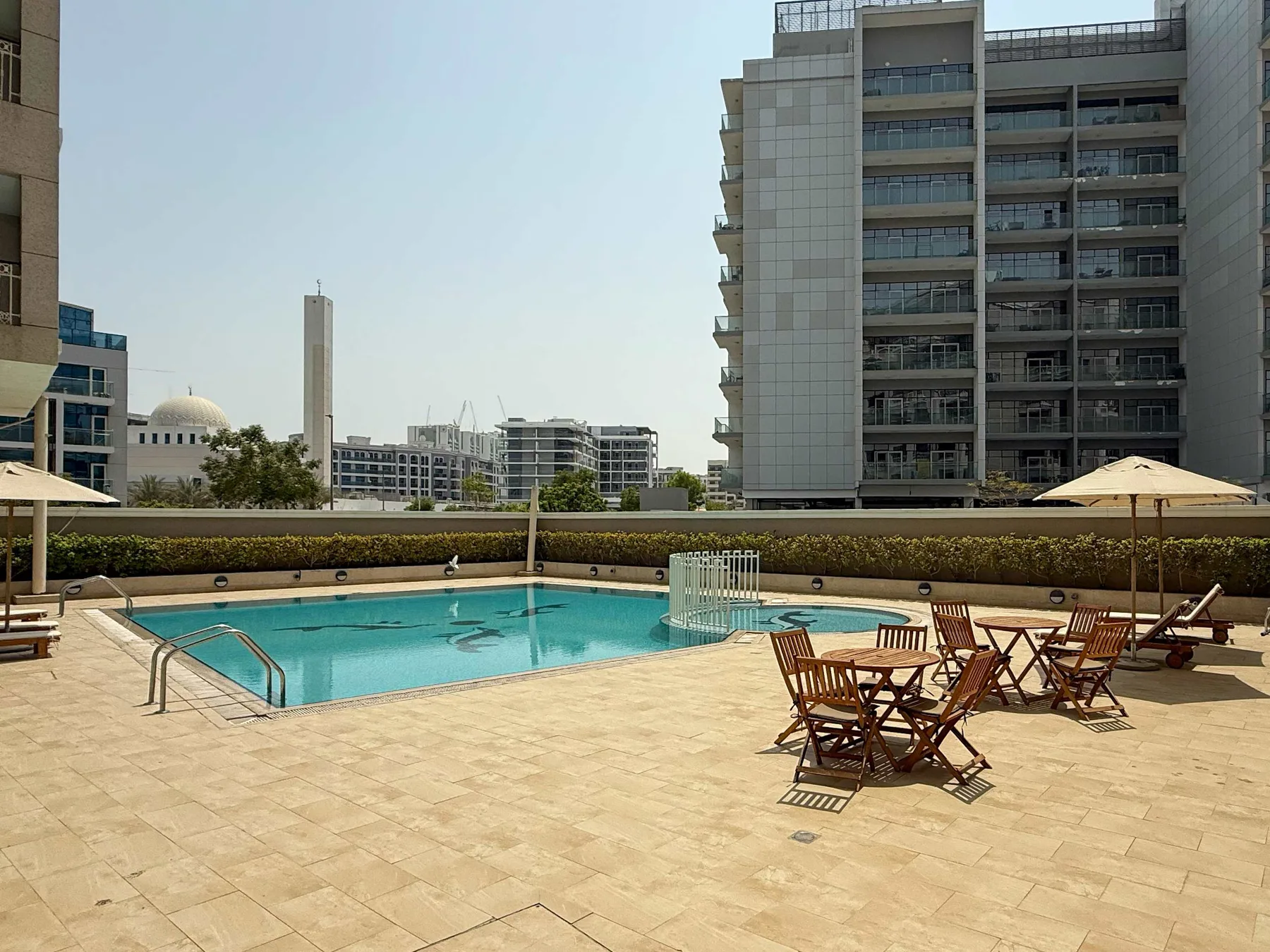 An outdoor pool and paved terrace with tables and seating beside apartment buildings.