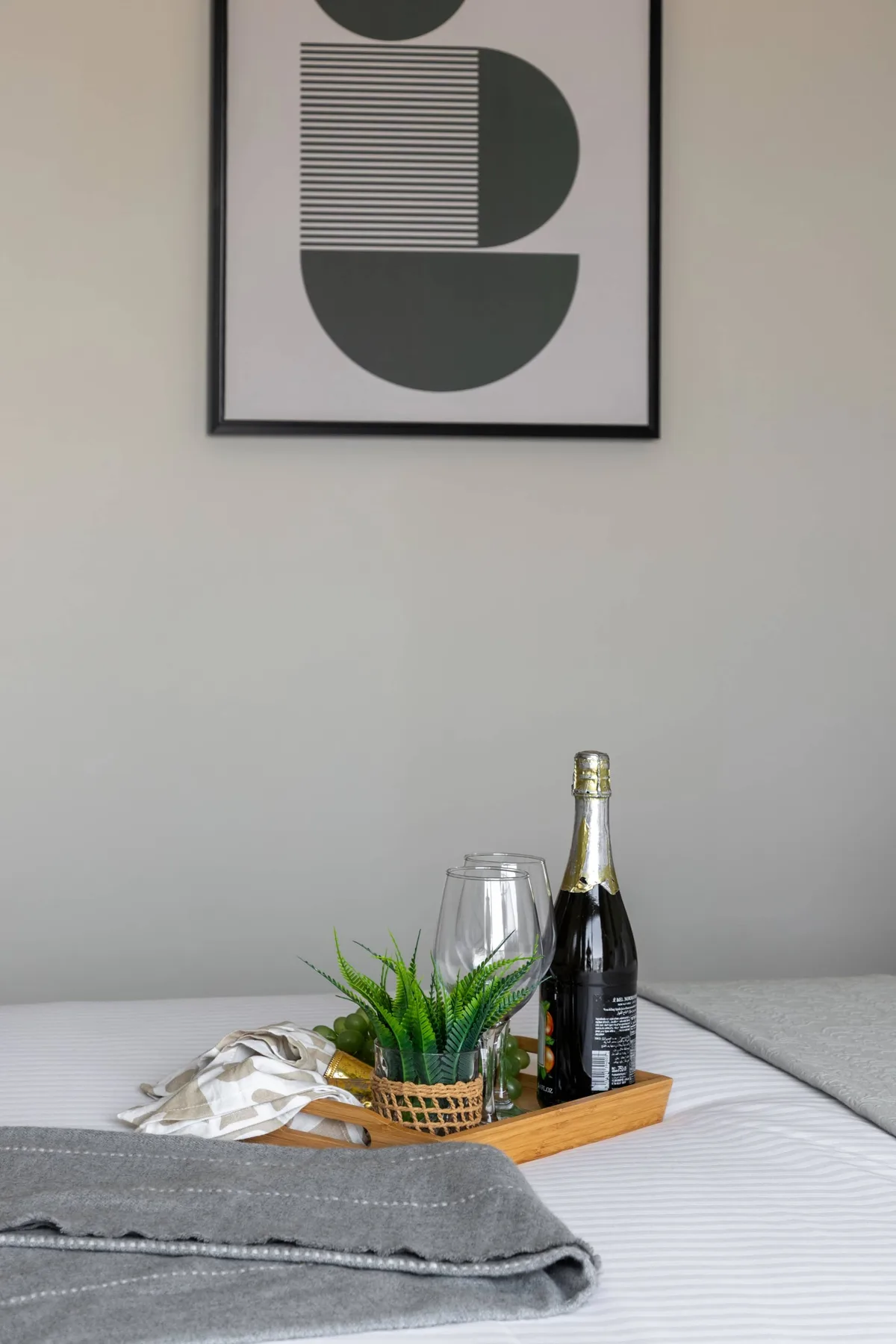 A neatly made bed displays a tray with glasses, a bottle, and a small plant.