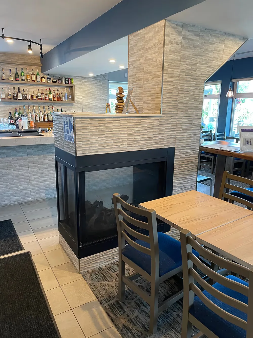 Zona de comedor con mesas de madera y sillas azules junto a una chimenea con panel de vidrio y un bar.