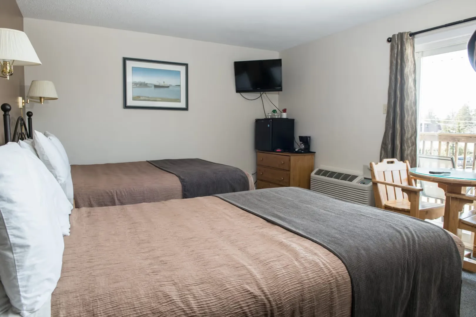 Habitación de hotel con dos camas dobles, televisor de pared, refrigerador pequeño y mesa con sillas de madera cerca de una puerta corrediza.