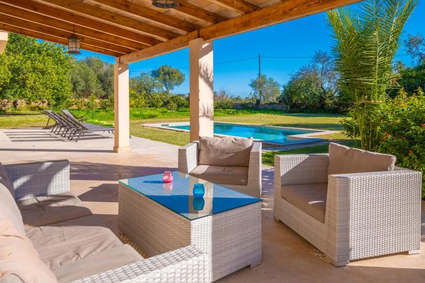 Un patio cubierto tiene asientos de mimbre alrededor de una mesa con tapa de vidrio, frente a una piscina.