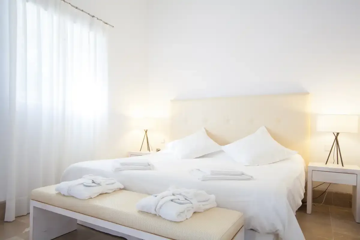 Un dormitorio luminoso cuenta con una cama con ropa blanca y un banco.