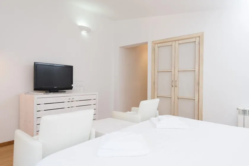 Un dormitorio luminoso con ropa de cama blanca, un televisor y una cómoda clara.