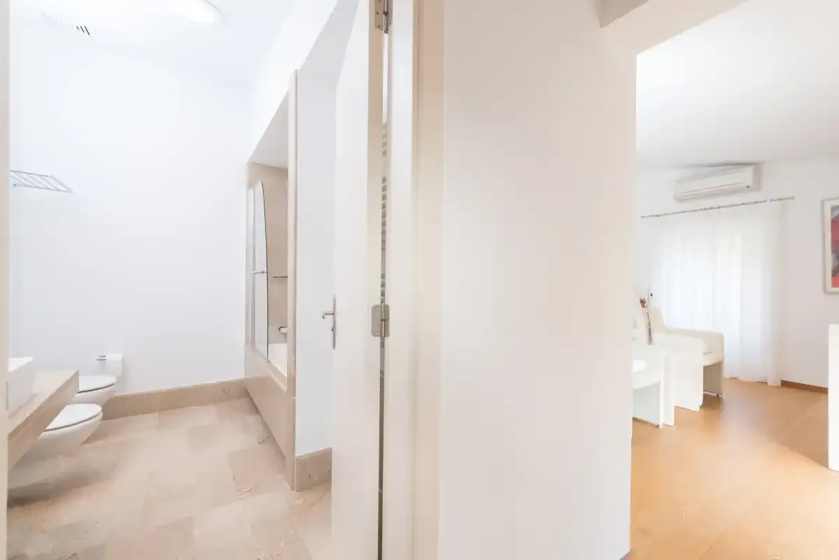 Un baño luminoso se abre a una habitación con escritorio y suelo de madera clara.
