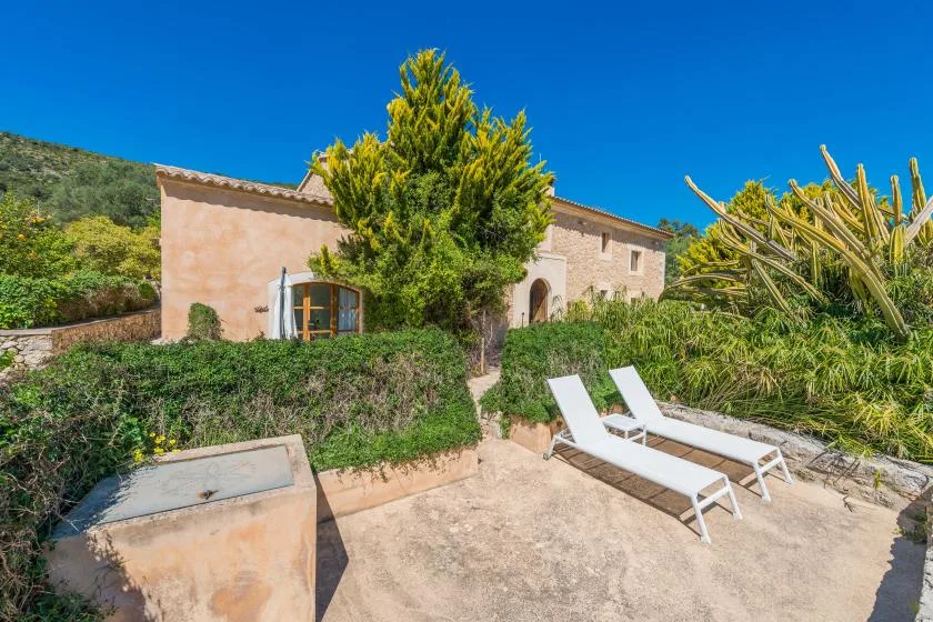Una casa de piedra se alza tras un jardín, con tumbonas blancas en un patio soleado.