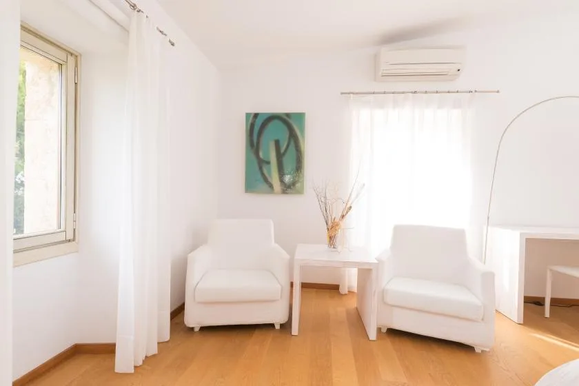 Una luminosa zona de estar tiene dos asientos blancos alrededor de una mesa pequeña, junto a grandes ventanas.