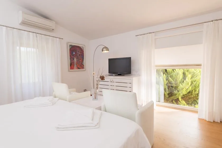 Un dormitorio luminoso con ropa de cama blanca, un televisor y grandes ventanas con cortinas.