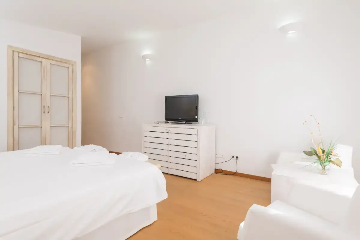 Un dormitorio luminoso con una cama blanca, una cómoda y un televisor.