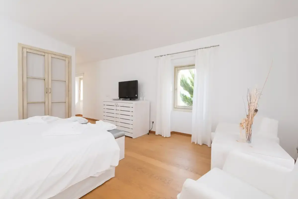 Un dormitorio luminoso con ropa de cama blanca, un televisor y una ventana.