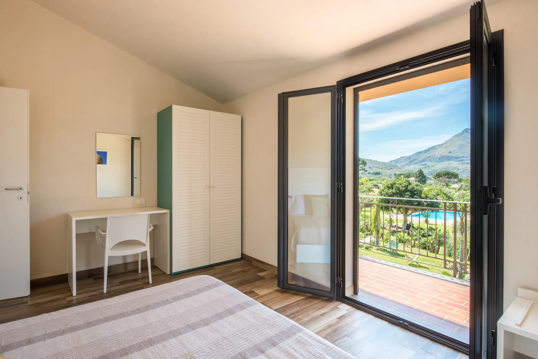 A bedroom with a desk and an open balcony door overlooking a green landscape.