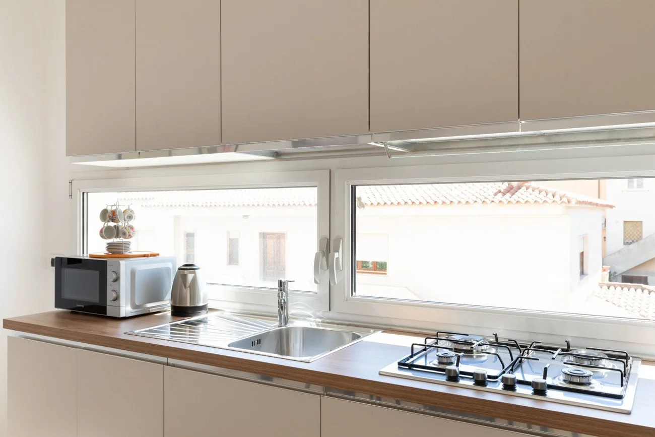 Una cocina luminosa con fregadero, microondas y cocina de gas bajo grandes ventanas.