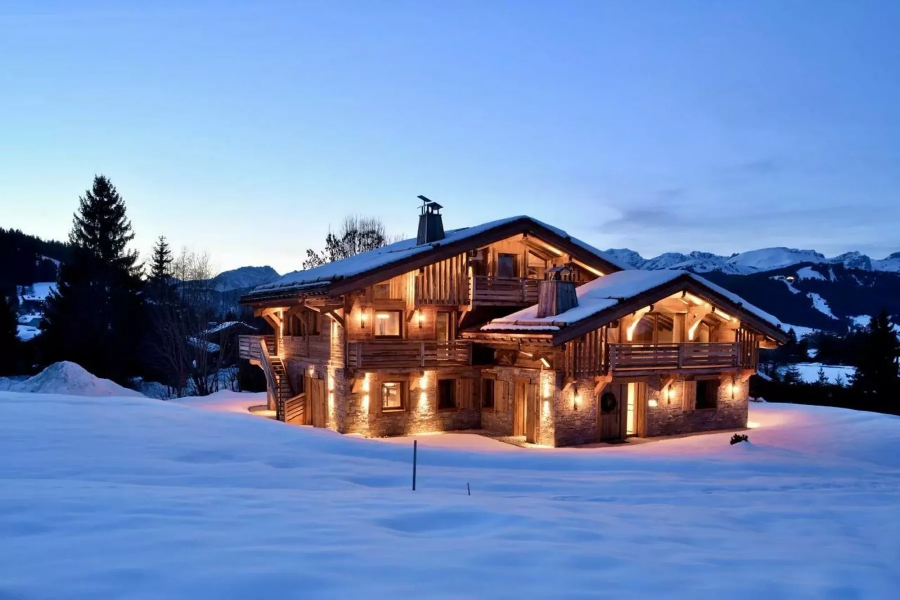 Un chalet de madera iluminado se alza en un paisaje nevado de montaña al anochecer.