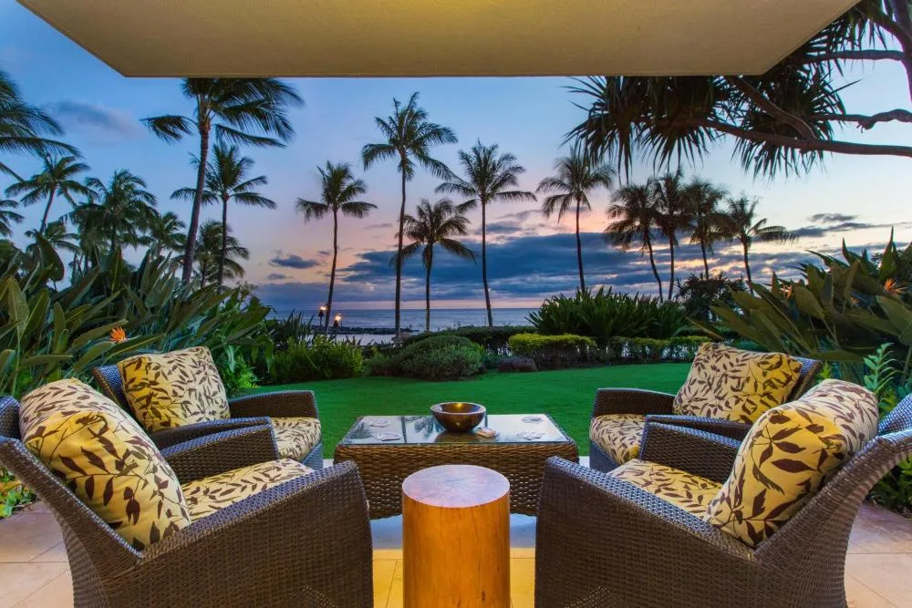 Un espacio de asientos al aire libre mira hacia un jardín, palmeras y el océano al atardecer.