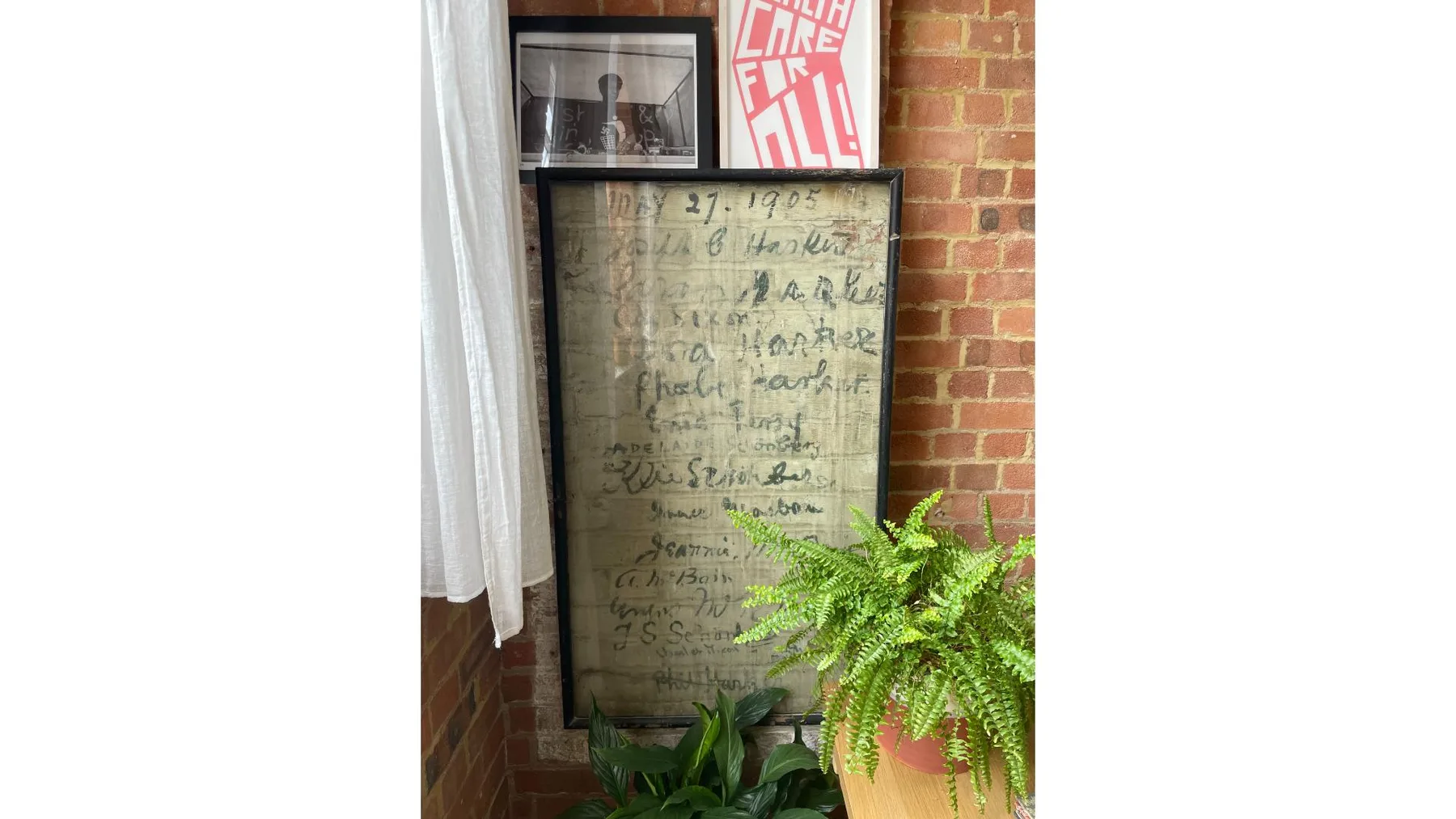 A brick-walled interior corner has framed artwork, a curtain, and leafy plants.