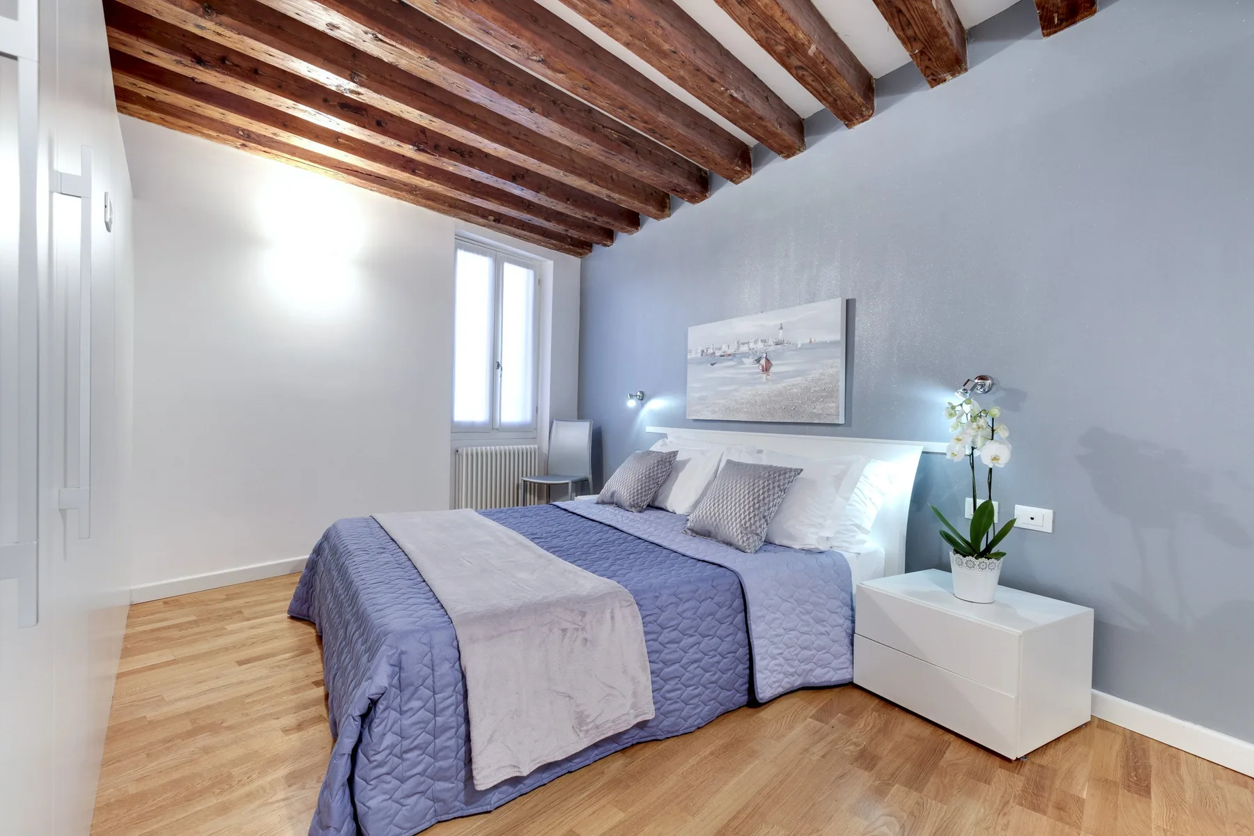 A bedroom with a double bed, pale bedding, and exposed wooden ceiling beams.