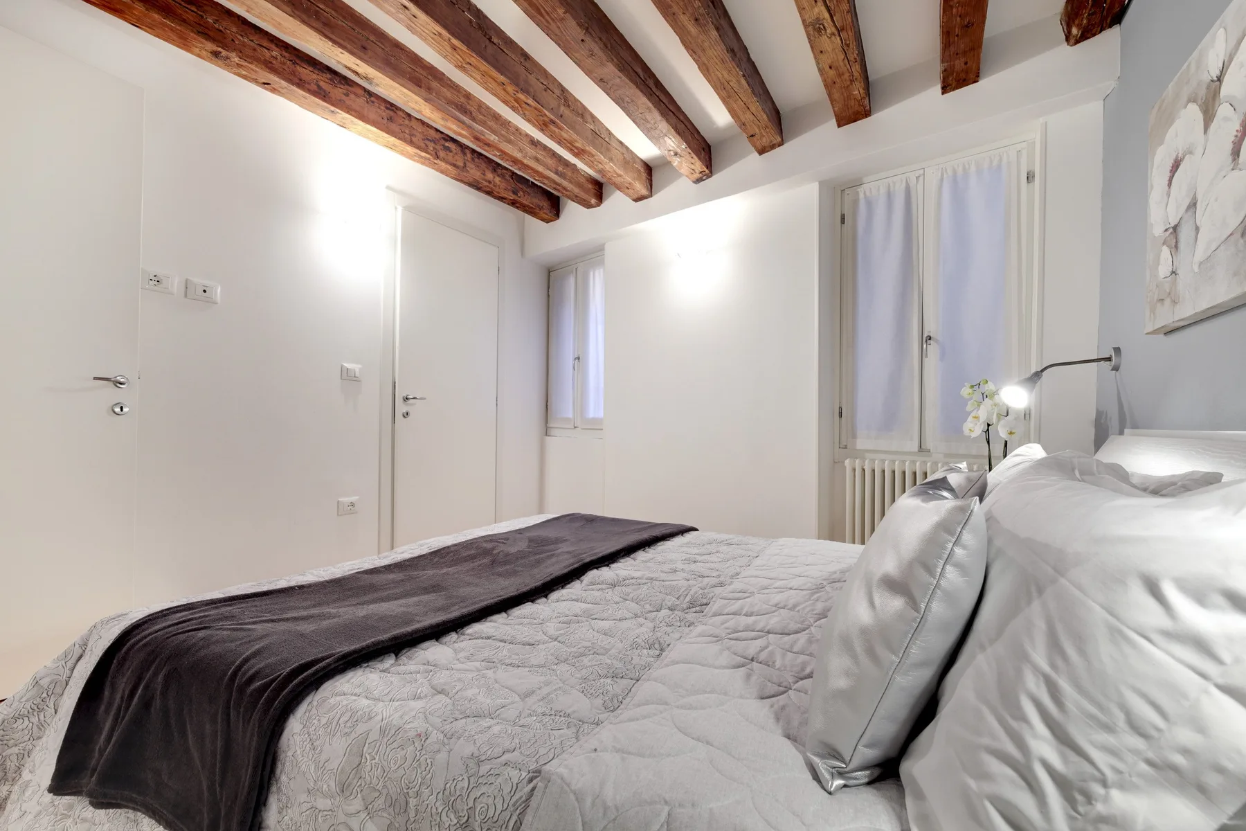 A bright bedroom with a neatly made bed beneath exposed wooden ceiling beams.