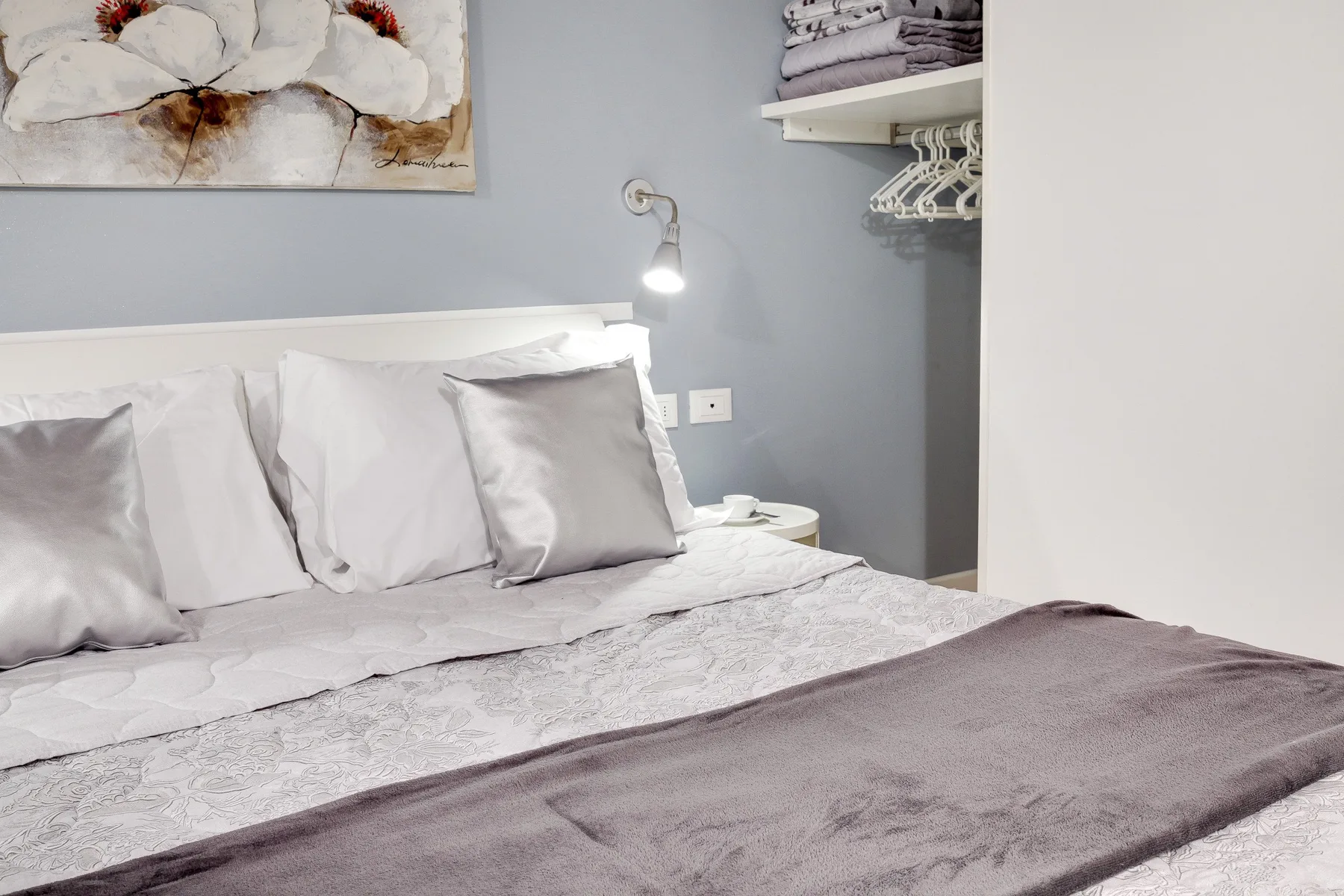 A neatly made bed with layered bedding sits beside a wall-mounted reading light.