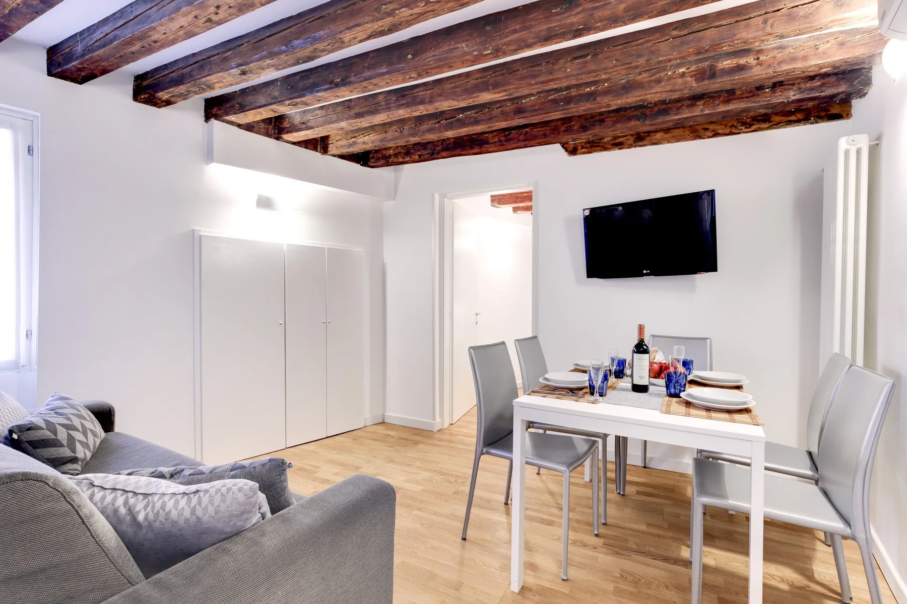 A bright dining and living area with a white table and gray seating beneath exposed wooden beams.