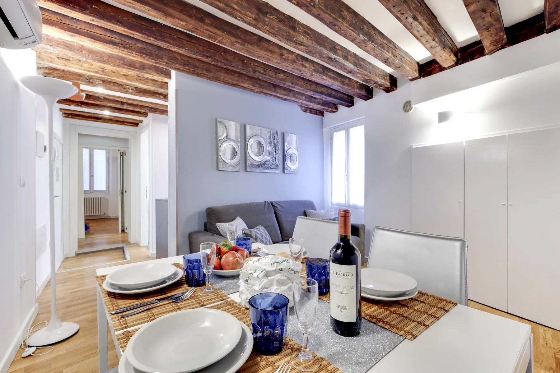 A dining table set with plates and glasses sits beside a living area with gray seating.