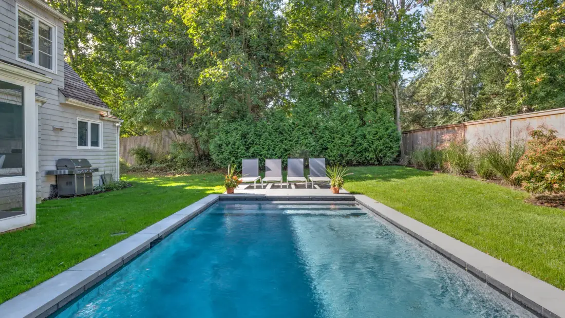 Una piscina en el patio está rodeada de césped verde y árboles frondosos.