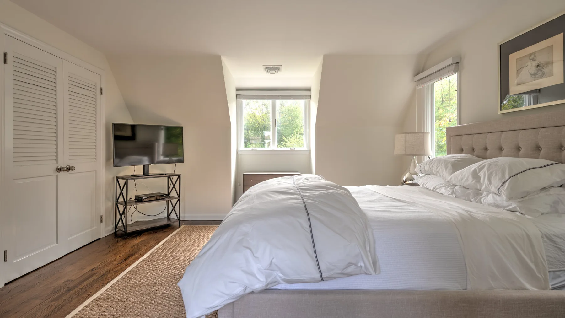 Un dormitorio luminoso con una cama junto a las ventanas y un televisor cerca.