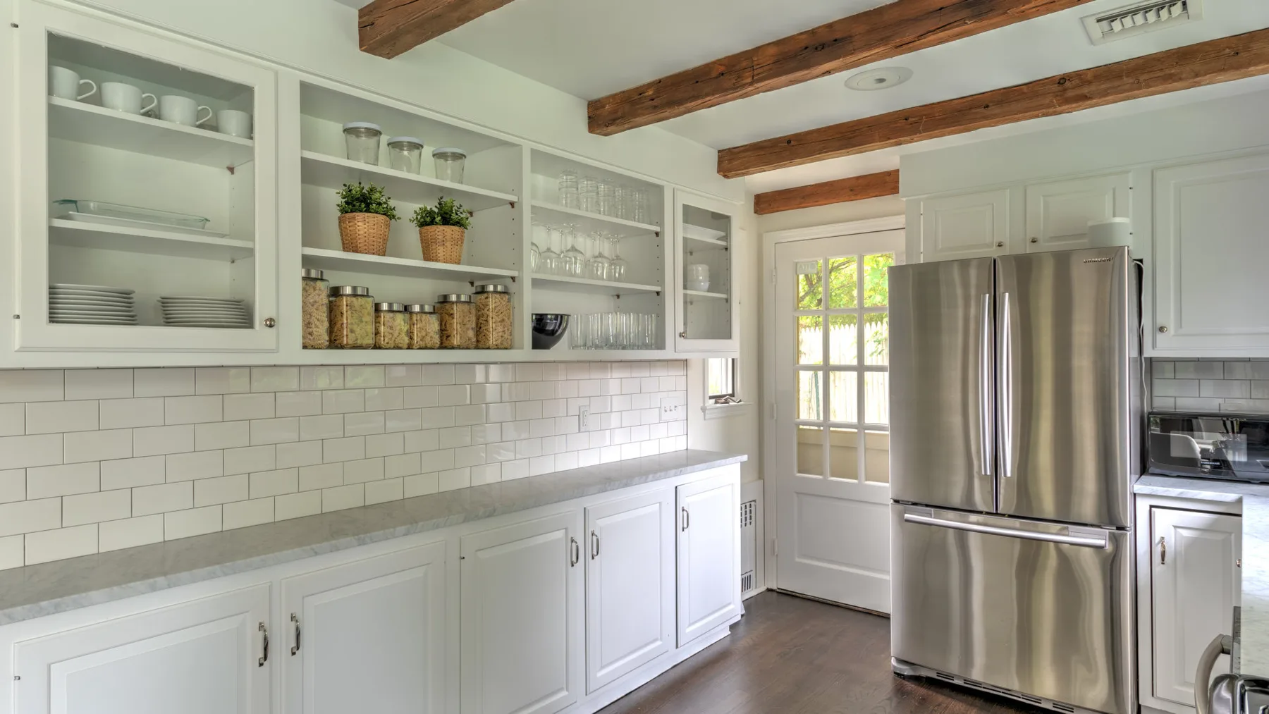 Una cocina luminosa con gabinetes blancos, estantes abiertos y un refrigerador de acero inoxidable.