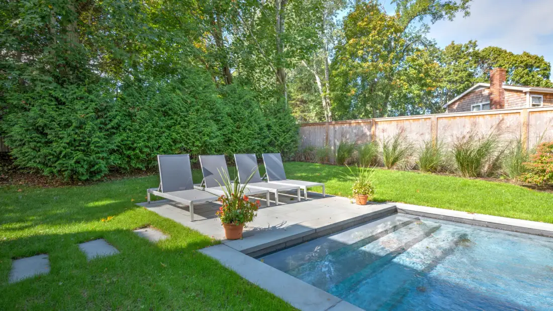Una piscina en el patio trasero está junto a una terraza pavimentada con asientos al aire libre.