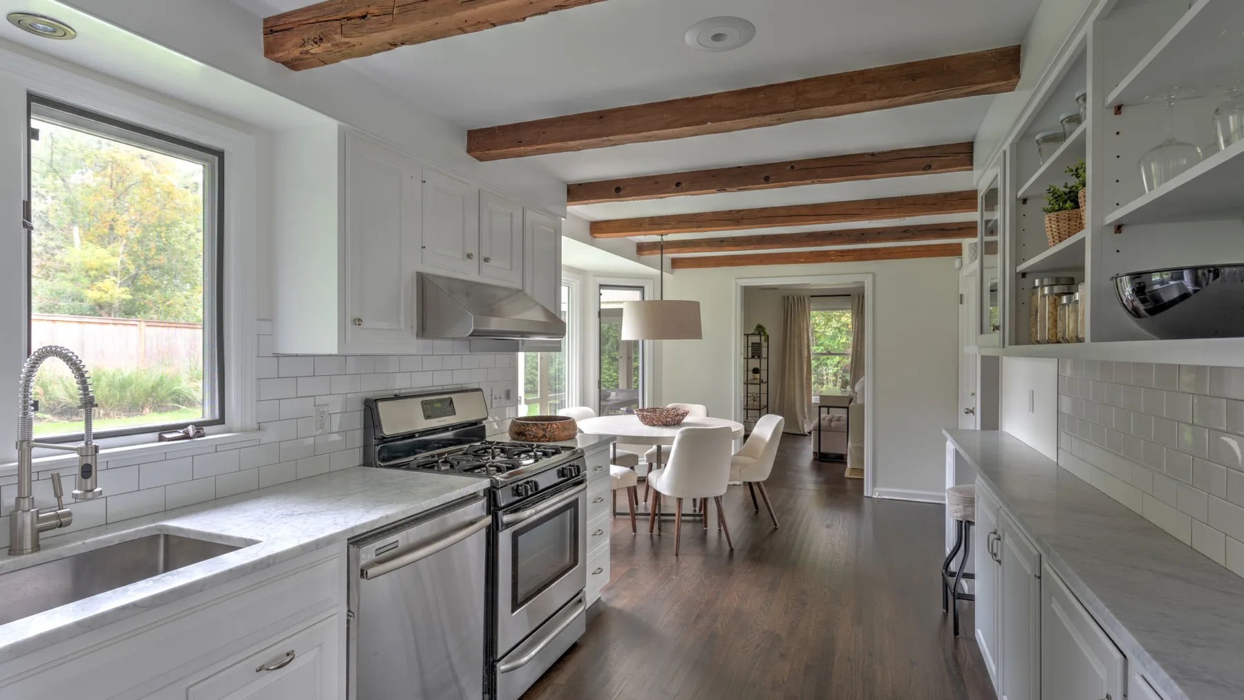 Una cocina luminosa se abre a un comedor, con gabinetes blancos y vigas expuestas en el techo.