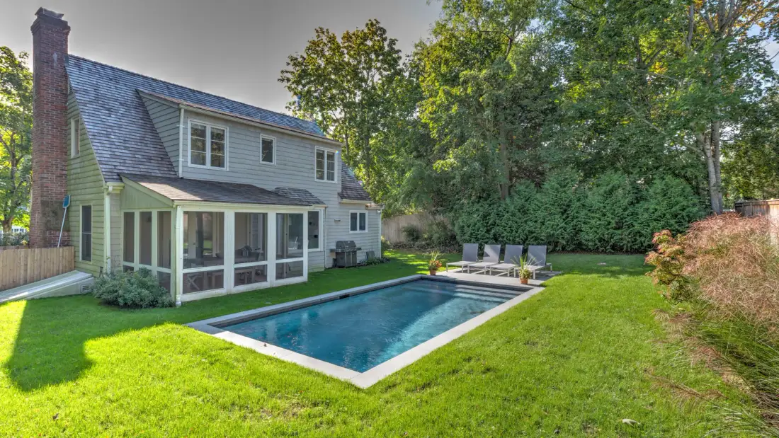 Una casa y una piscina rectangular están rodeadas de césped verde y árboles.