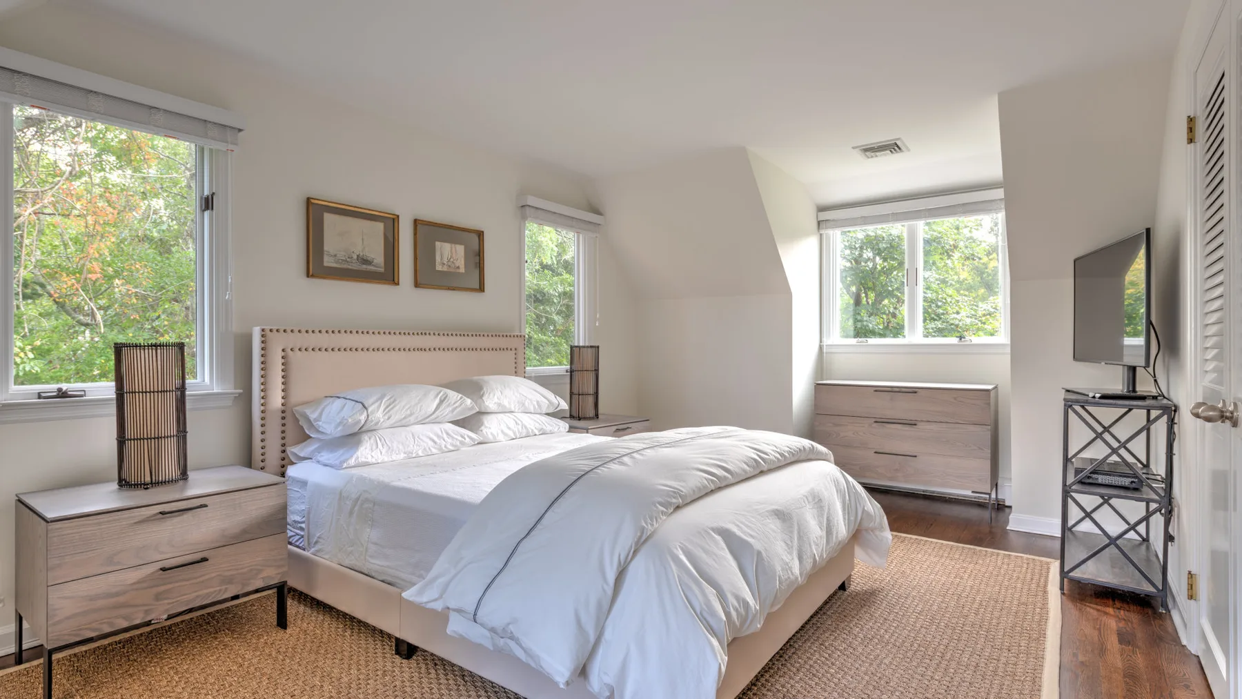 Un dormitorio luminoso con una cama tendida y ventanas con vistas a la vegetación.