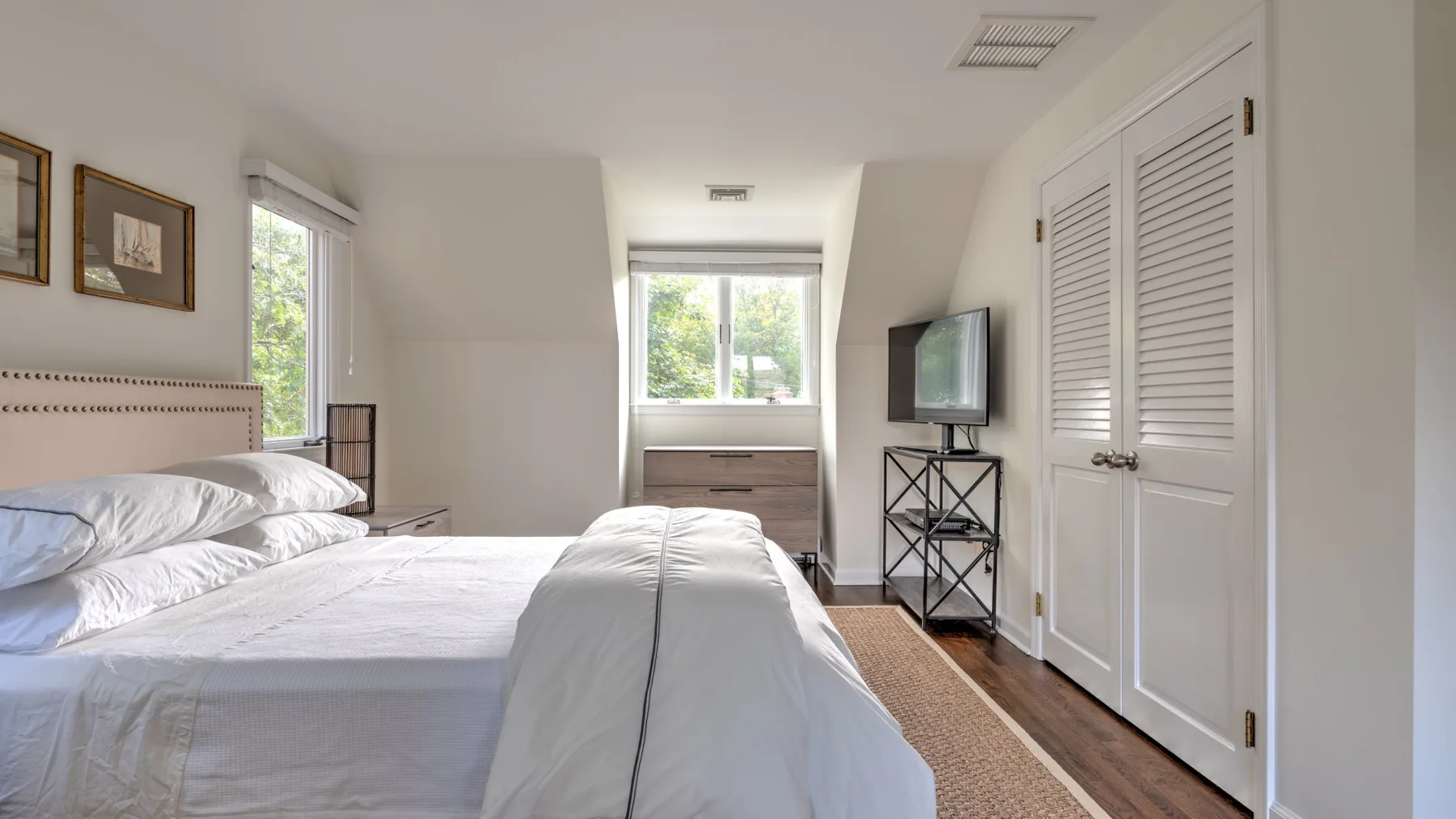 Un dormitorio luminoso con una cama bien hecha, ventanas y un televisor sobre un soporte.