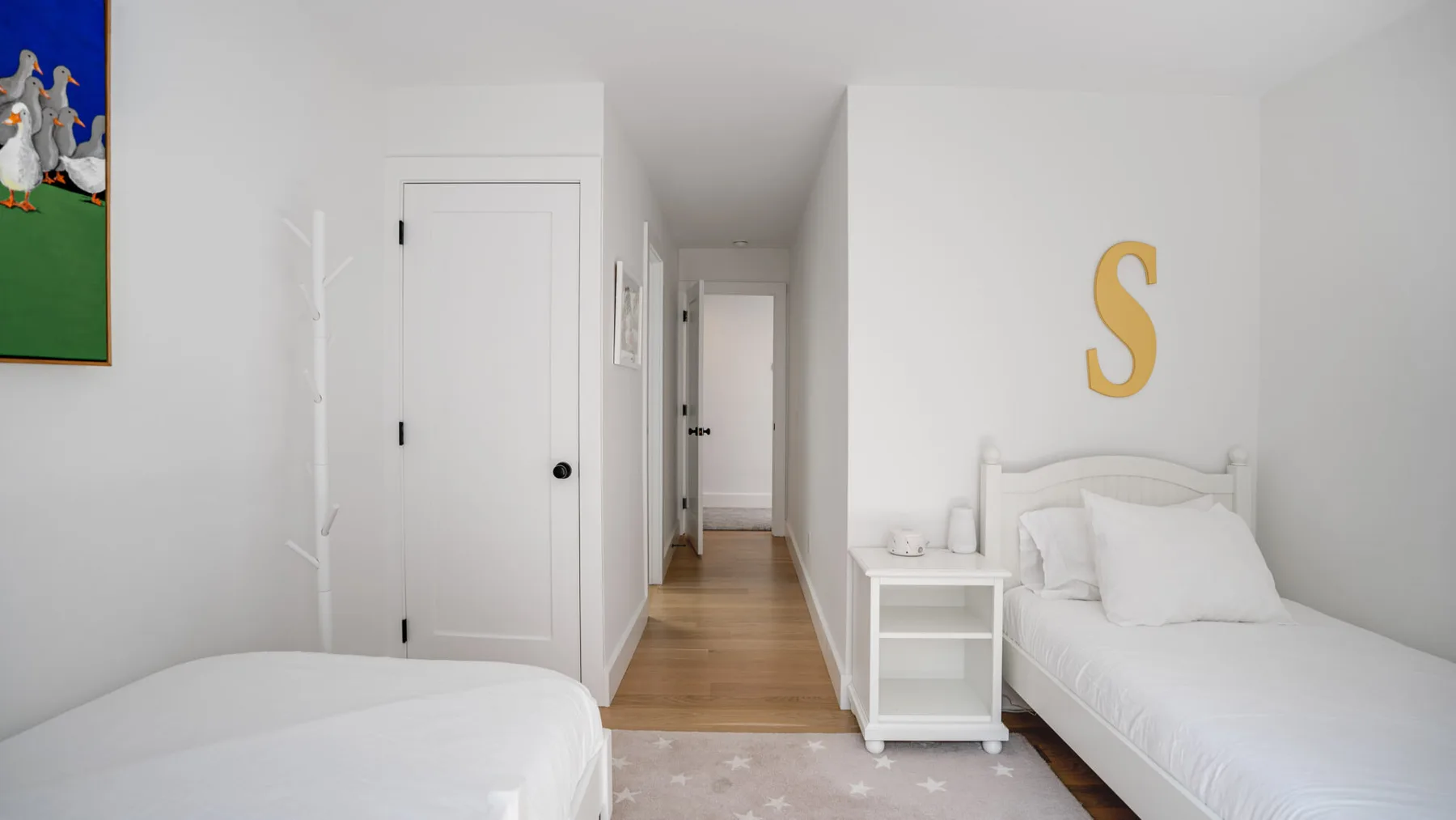 Un dormitorio luminoso con camas blancas, una mesita de noche y un pasillo.